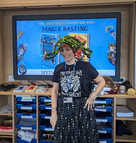 Marie Basting Children's Author wearing a silly shiny green Medusa-inspired snake hat in a classroom with author visit props in the background.