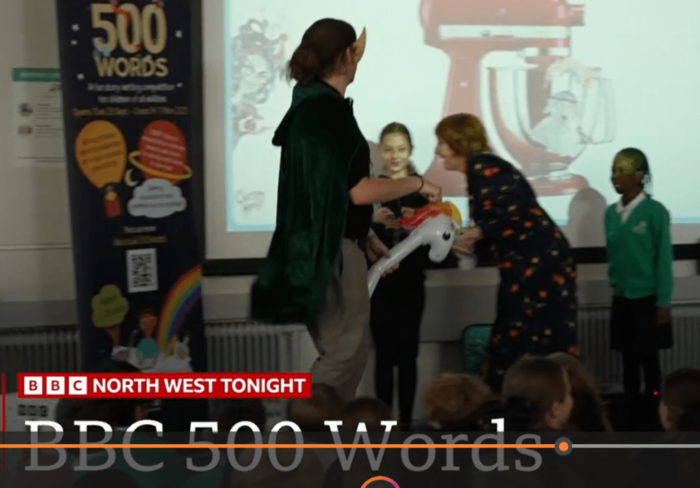 Teacher dressed in green cape and elf ears rides hobby horse unicorn while Marie Basting author and school children from Blackley Academy share a laugh